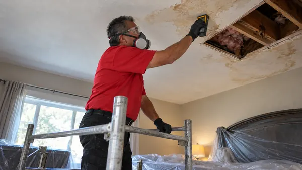 Technician in a respirator reaching up to read moisture on a stained ceiling beside an open attic access hole