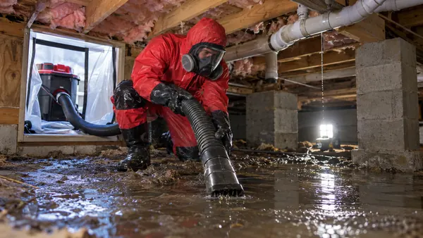 Worker in a hooded red suit and respirator pumping muddy black water out of a flooded crawl space past block piers and pink insulation